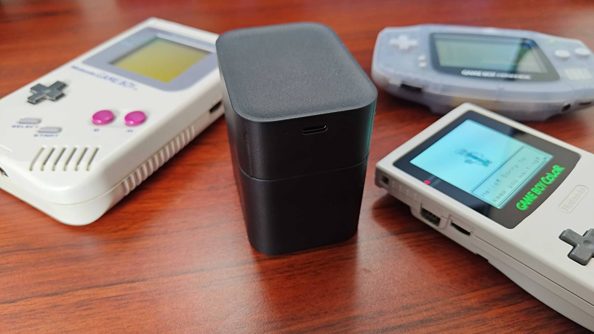 Rechargeable battery case surrounded by Game Boy, Game Boy Color, and Game Boy Advance SP on woodgrain desk.