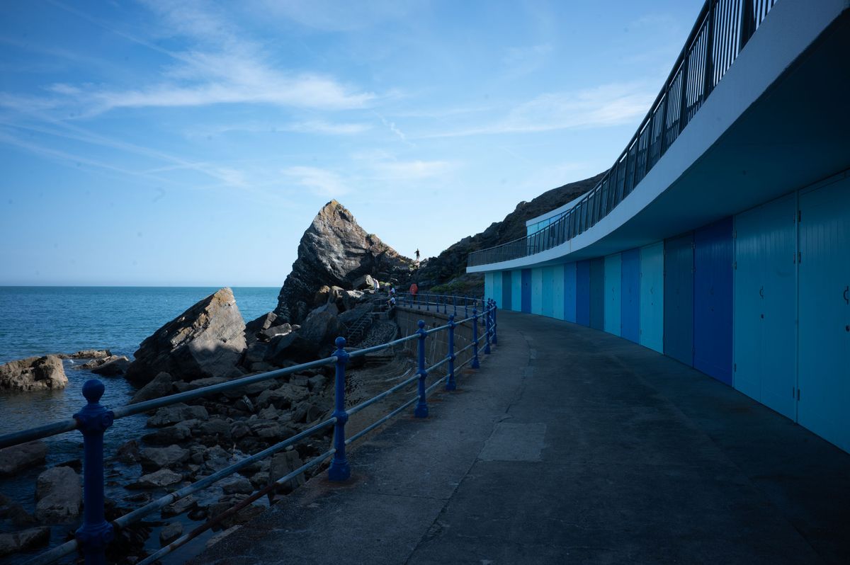 Blue beach huts arch around a concrete walkway, leading to a jagged rockside
