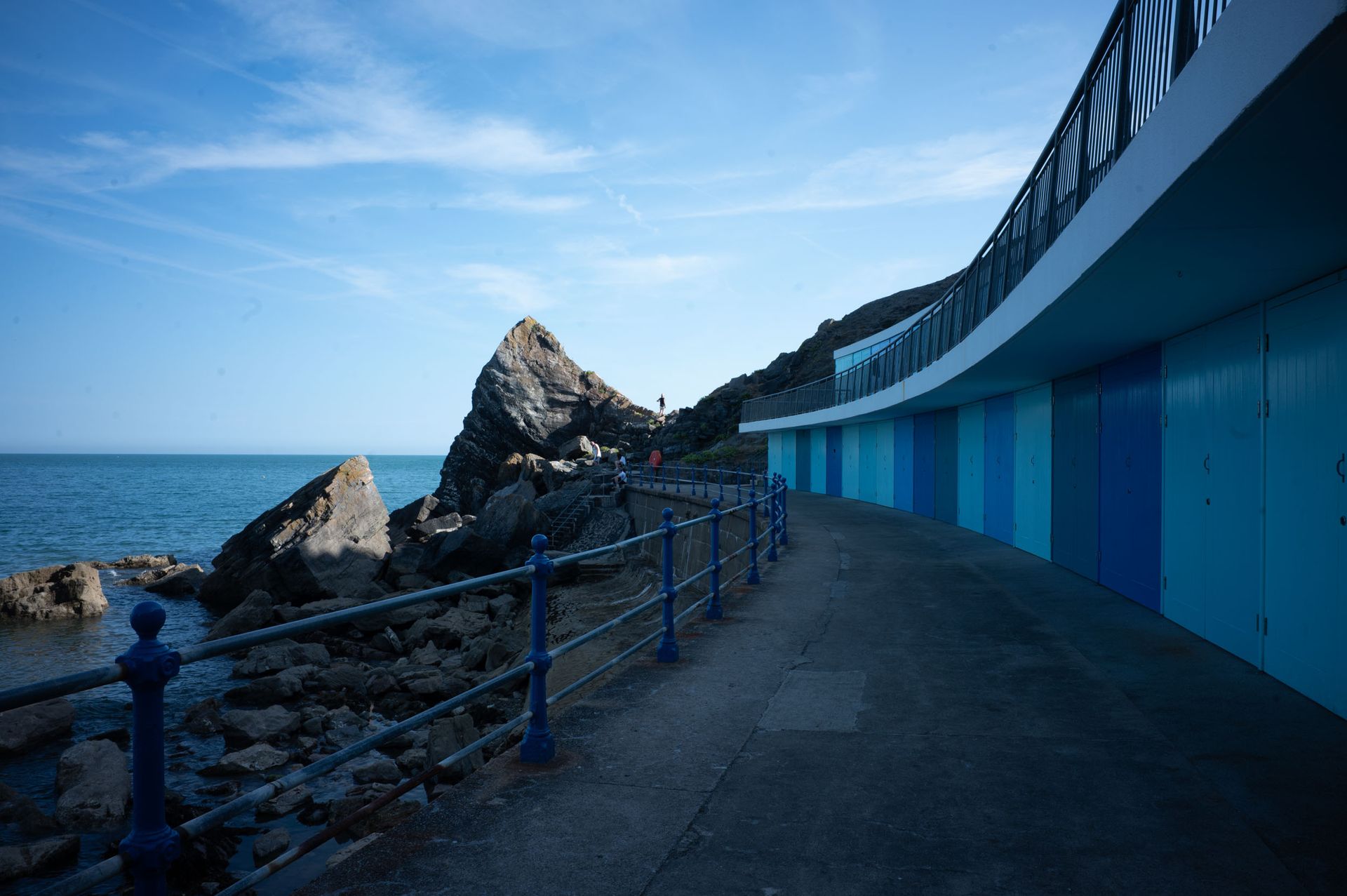 Blue beach huts arch around a concrete walkway, leading to a jagged rockside