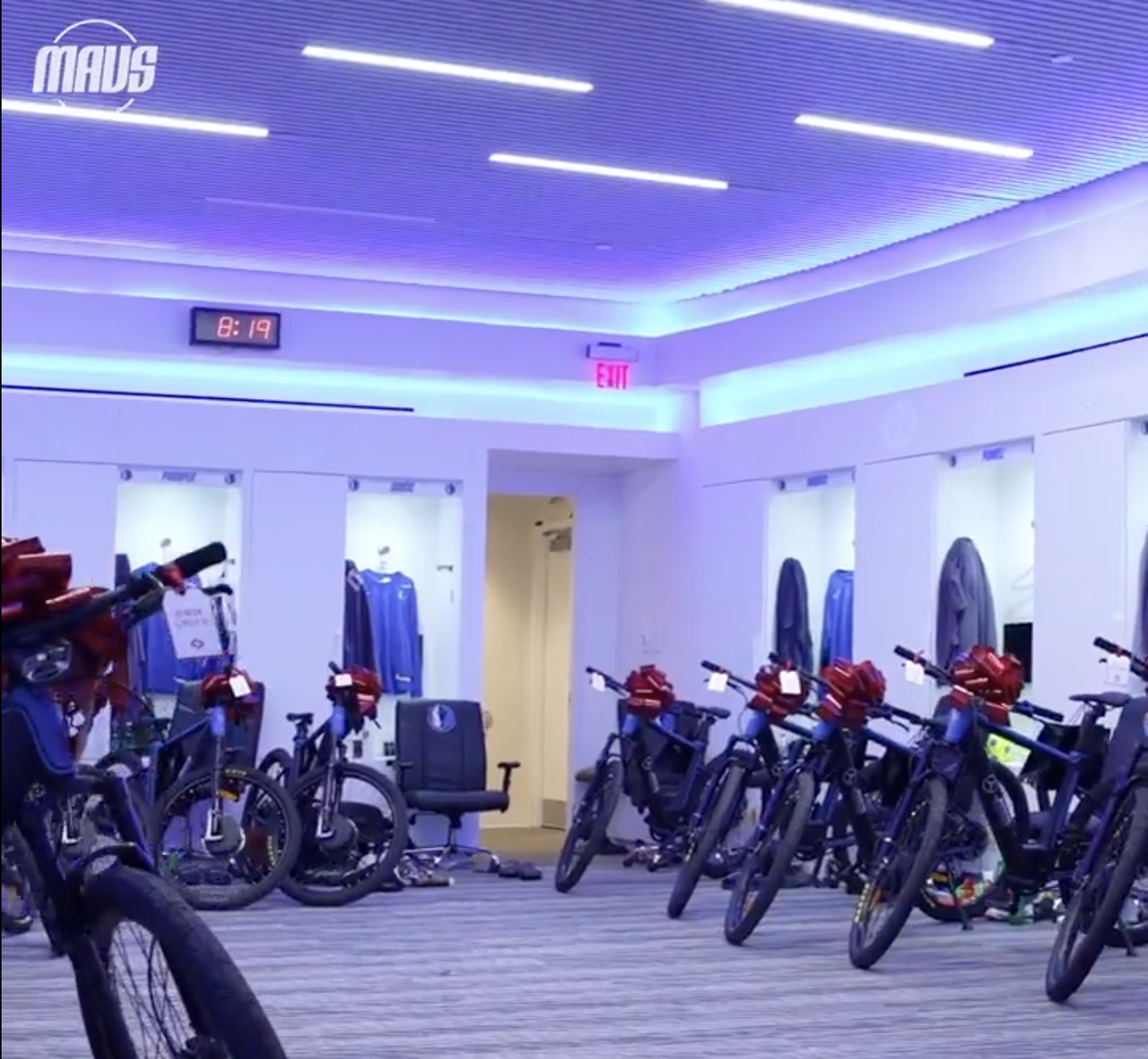 A row of black e-bikes with big red bows on their flat handlebars are lined up in a blue-lit carpeted basketball locker room.