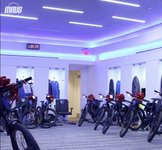 A row of black e-bikes with big red bows on their handlebars are lined up in a blue-lit carpeted basketball locker room.