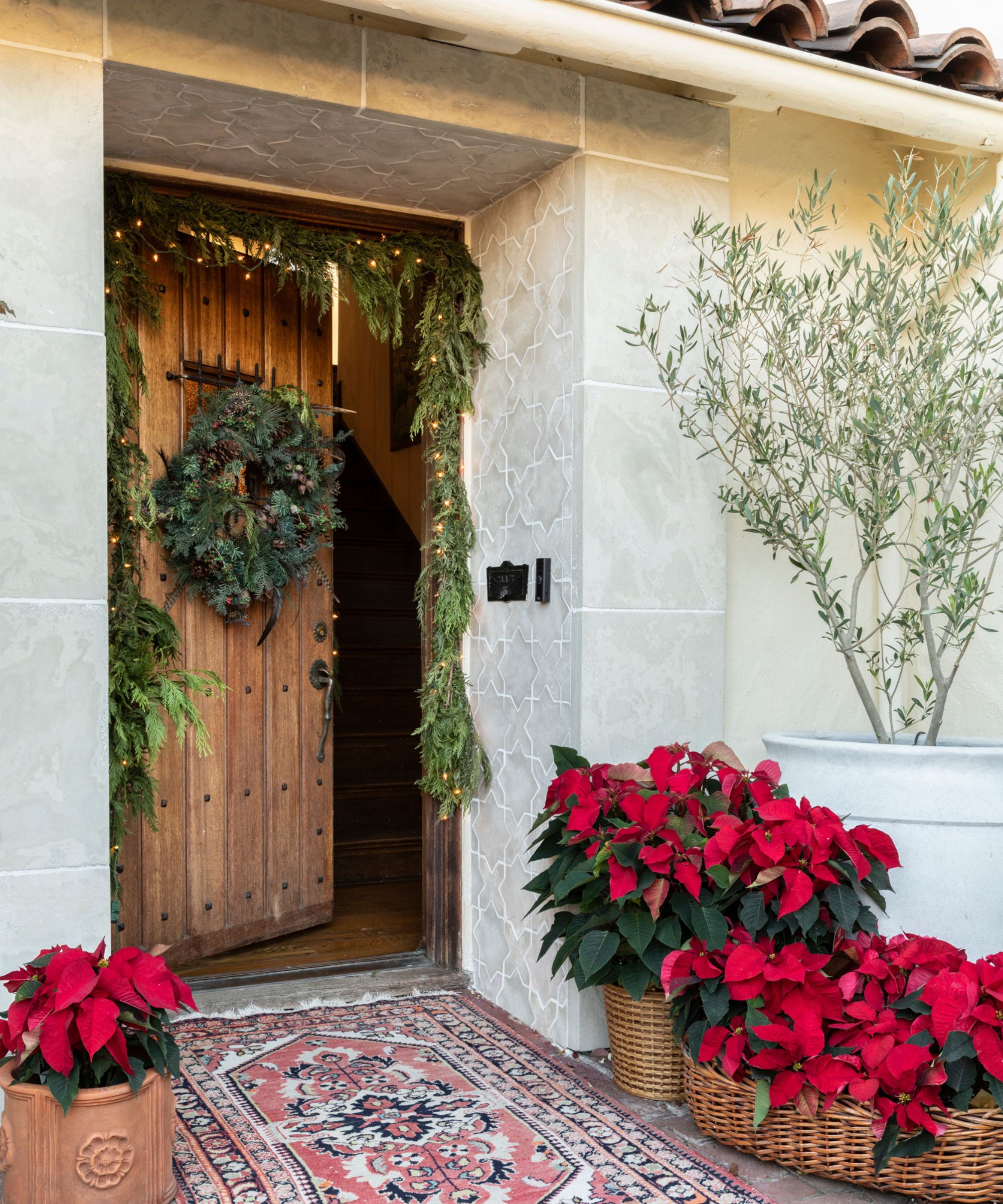 A wooden front door decorated with a natural wreath and garland, both wrapped in warm lights. In front, woven baskets have been filled with classic red poinsettia plants