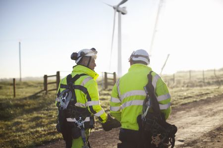 Two engineers in safety garb walk by a windfarm.