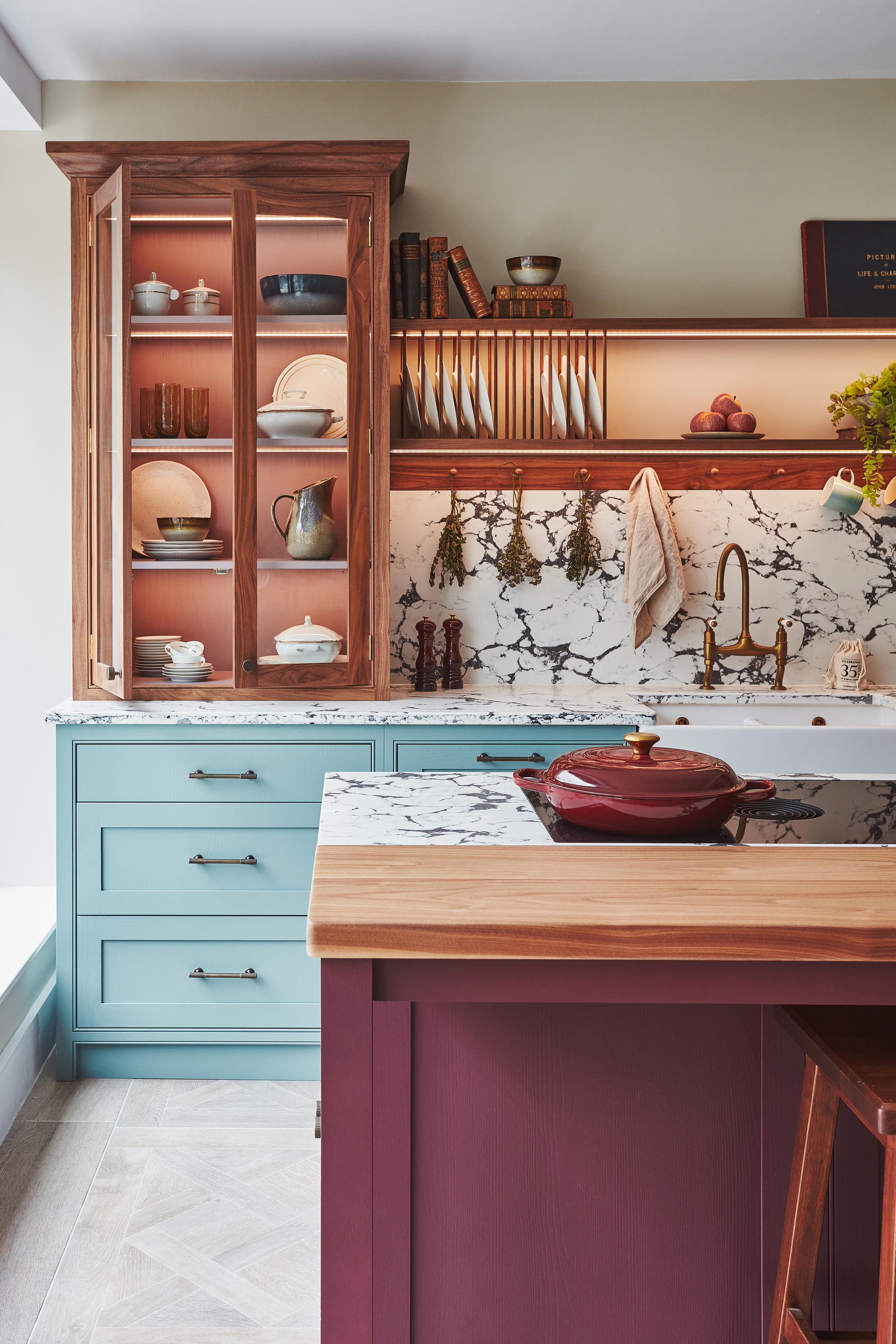colorful kitchen with light blue and plum cabinets, with exposed timber and brass accents, marble countertop and backsplash, and gray herringbone floors