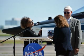 Johnson Space Center director Vanessa Wyche and former center director Michael Coats present Susy Young with a plaque honoring her late husband and the naming of Hangar 276.