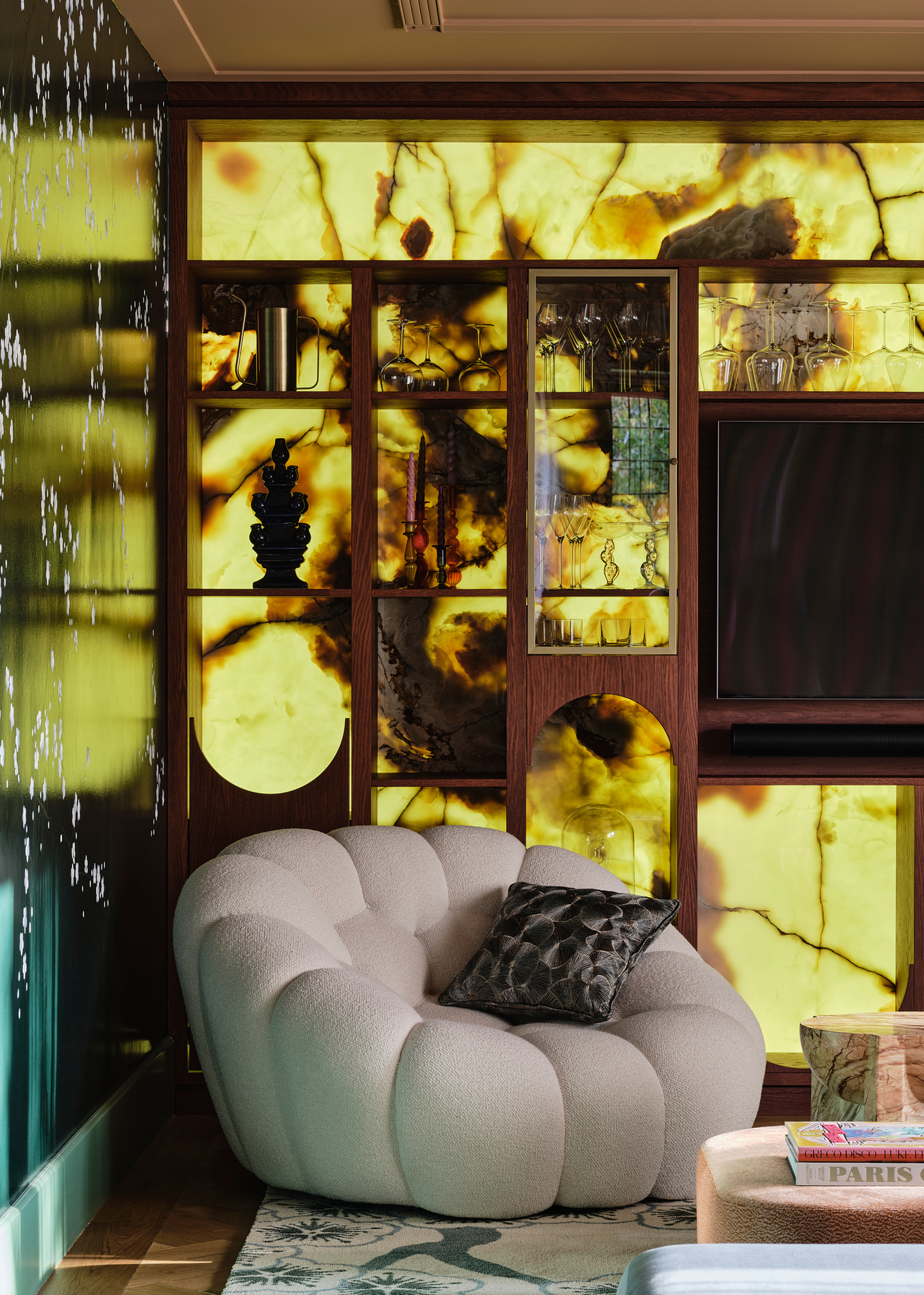a corner of a living room with a backlit display cabinet and a shell-like cream armchair with a cushion in the corner