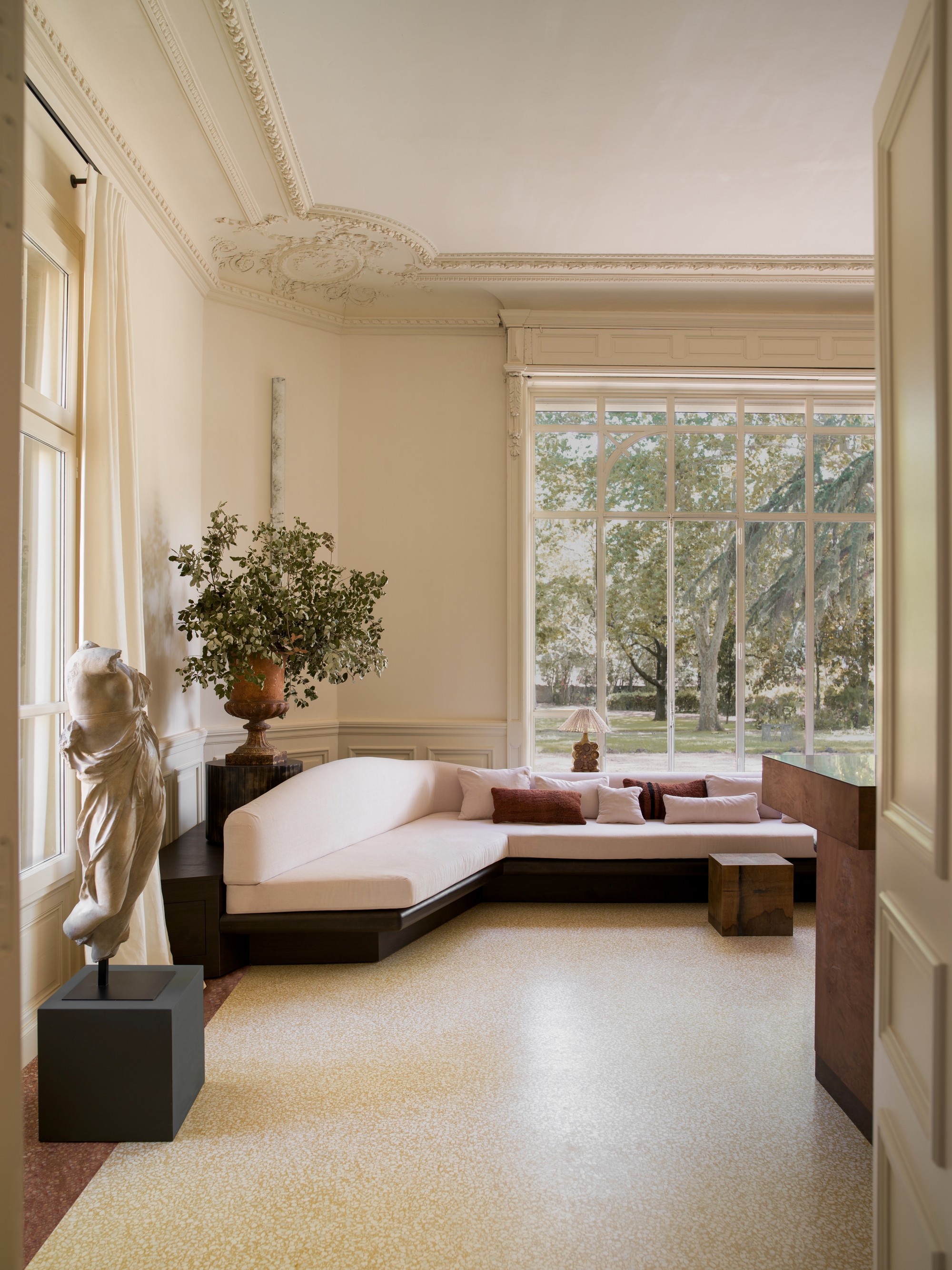 A grand, living room with large window and intricate crown molding. There are yellow terrazzo floors and a large black sectional sofa with light pink cushions.