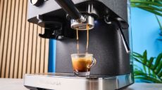 a black espresso machine by kitchenaid photographed against a blue background with silver chrome buttons and a pressure dial and steam wand