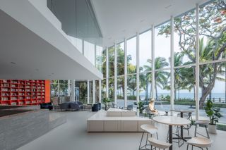 White interior, grey gloss marble counter, seating area, planters, tables and chairs, orange shelving unit on the left hand side wall, window framed viewing wall looking out to the surrounding landscape, trees, fencing and sea view, woman sat at a table looking out of the window, blue sky