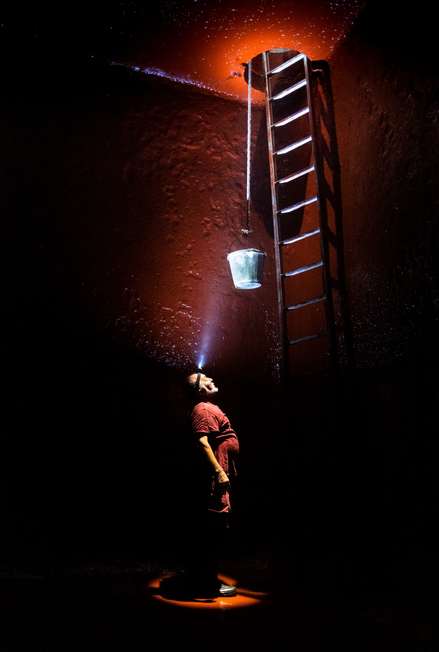 Man cleaning inside of empty wine vat looks up towards entrance of the vessel at the top of a ladder. A bucket hands via a rope from the entrance above.