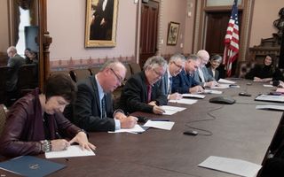 people in suits sit at a long table signing pieces of paper