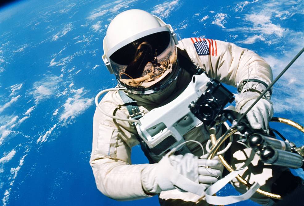 Astronaut Ed White floats in the microgravity of space outside the Gemini IV spacecraft. Behind him is the brilliant blue Earth and its white cloud cover. White is wearing a specially-designed space suit. The visor of the helmet is gold plated to protect him against the unfiltered rays of the sun. In his left hand is a Hand-Held Self-Maneuvering Unit with which he controls his movements in space.