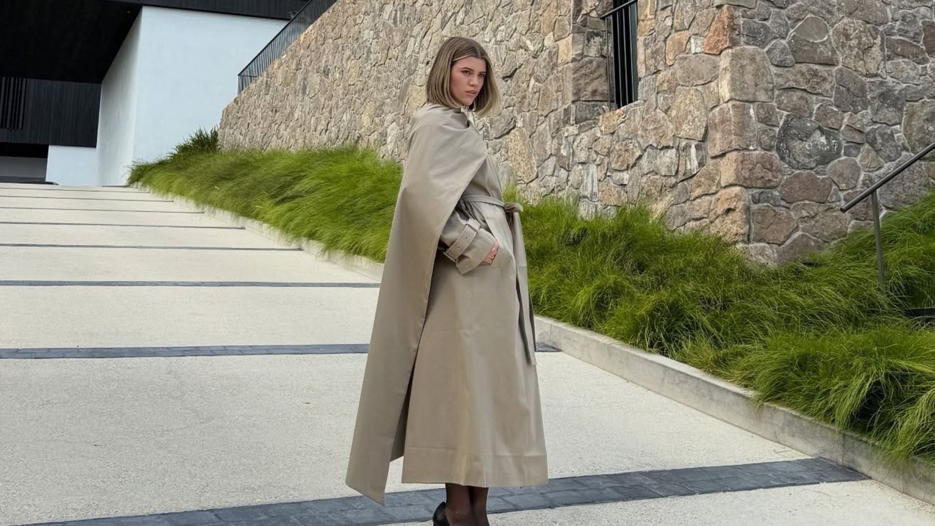 Sofia Richie poses for a photo outside wearing a long-line beige scarf coat with sheer tights and black pointed-toe heels.