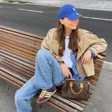 Fashion influencer Luna Isabella wearing a chic spring outfit styled with on-trend basics, including faded blue relaxed jeans, a white tee, a striped button-down shirt, a cropped trench, a baseball cap, a top-handle bowling bag, and low, vintage-inspired Adidas SL 72 OG sneakers.