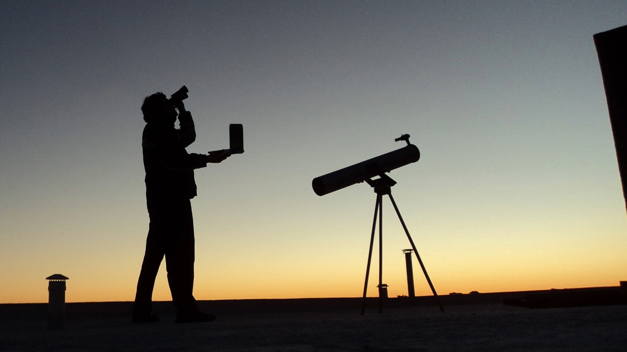 The silhouette of two people and their telescope are outlined in a glowing night sky