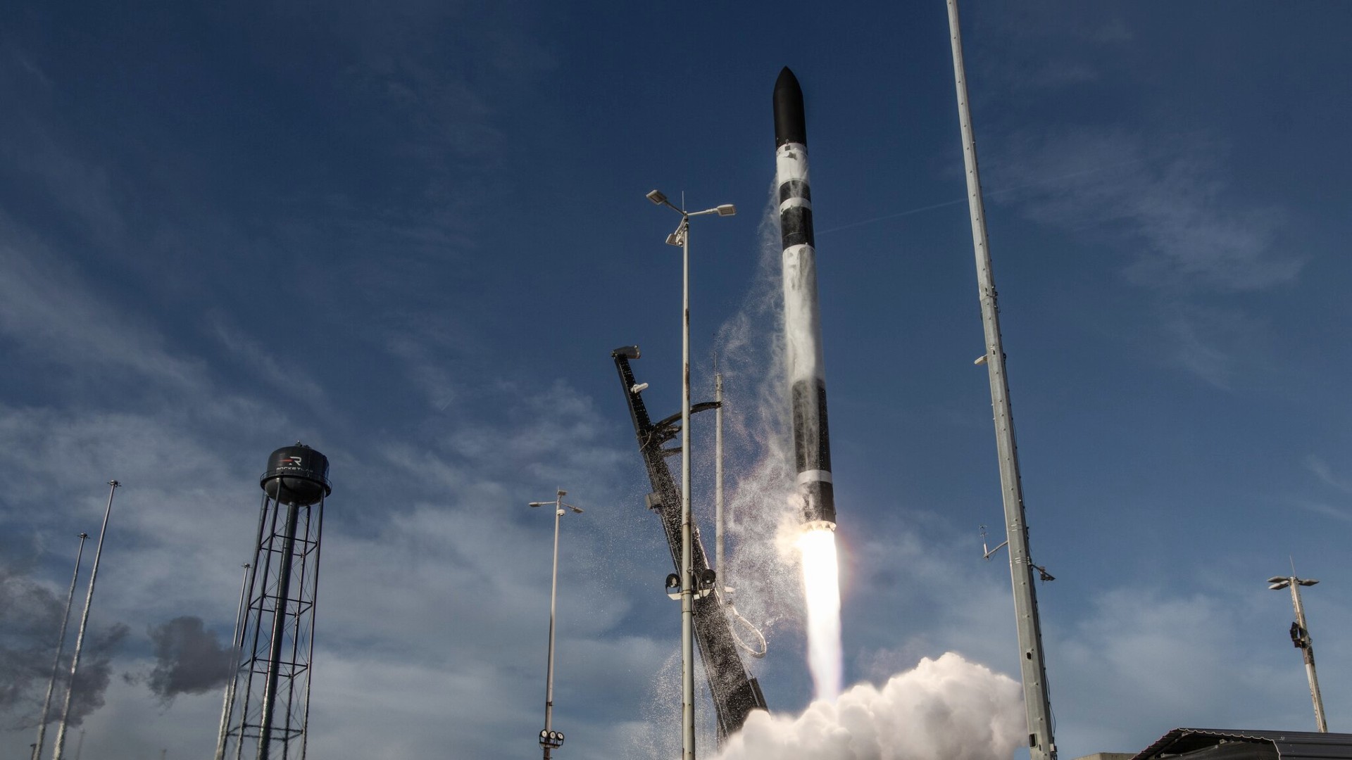 a black and white rocket lifts off above a cone of fire and smoke next two a black water tower