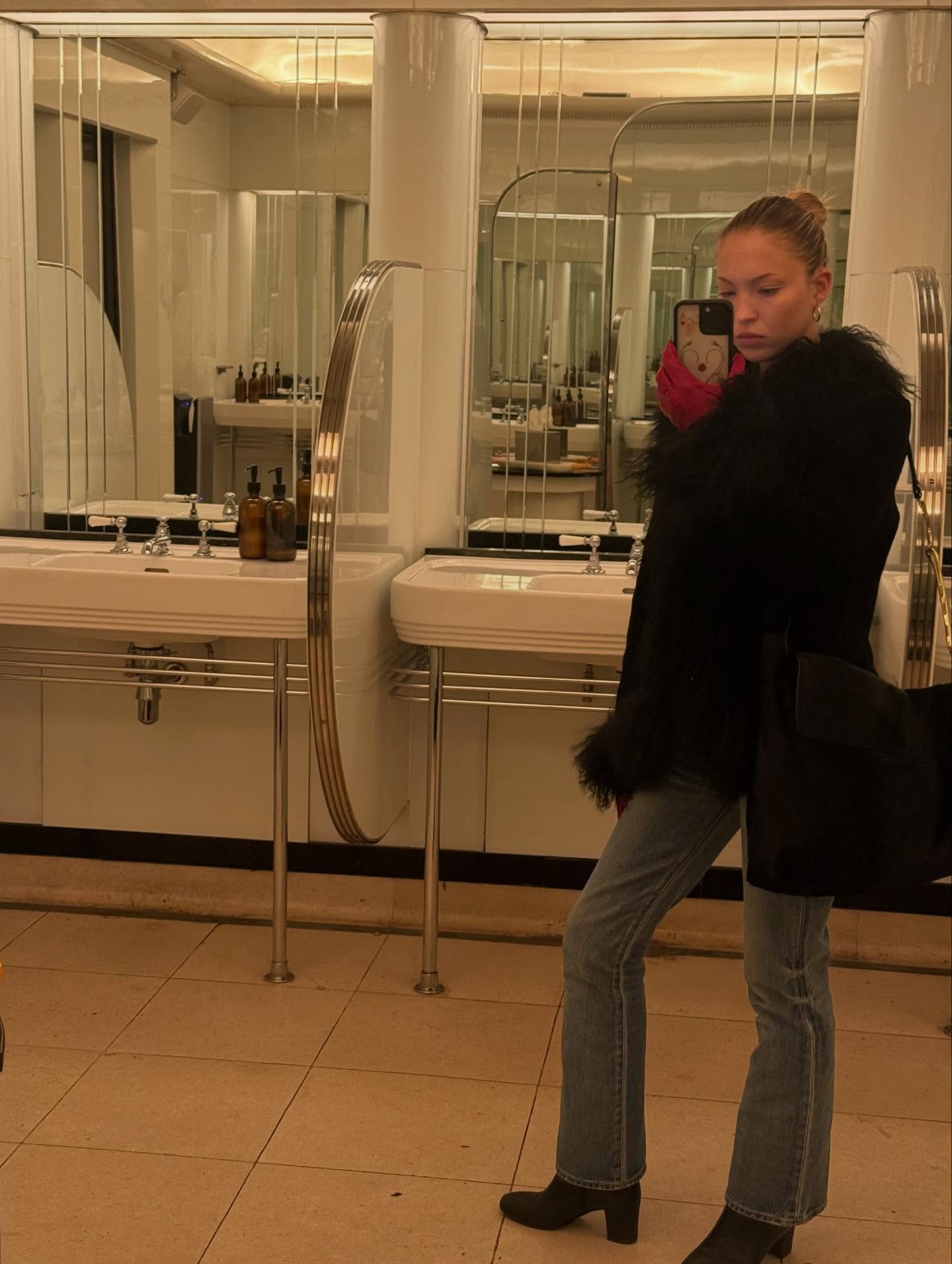 Lila Moss takes a mirror selfie in a bathroom wearing bootcut jeans with rounded-toe heels boots and a fur-trimmed jacket.