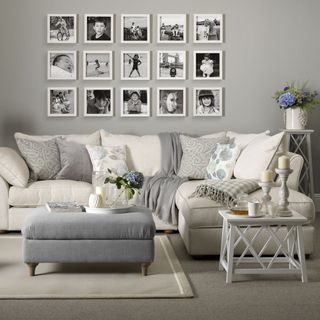 grey living room with wall-mounted picture frames with a cream-colored sofa with soft neutral, delicately patterned cushions on a grey carpet next to a white table topped with candles and tea