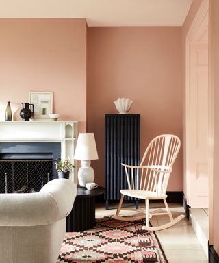 pastel living room ideas, plaster coloured pink walls with light shade on ceiling and doors, black skirtings and radiator, white floorboards, rocking chair, pattered rug, oatmeal couch, fireplace, artwork