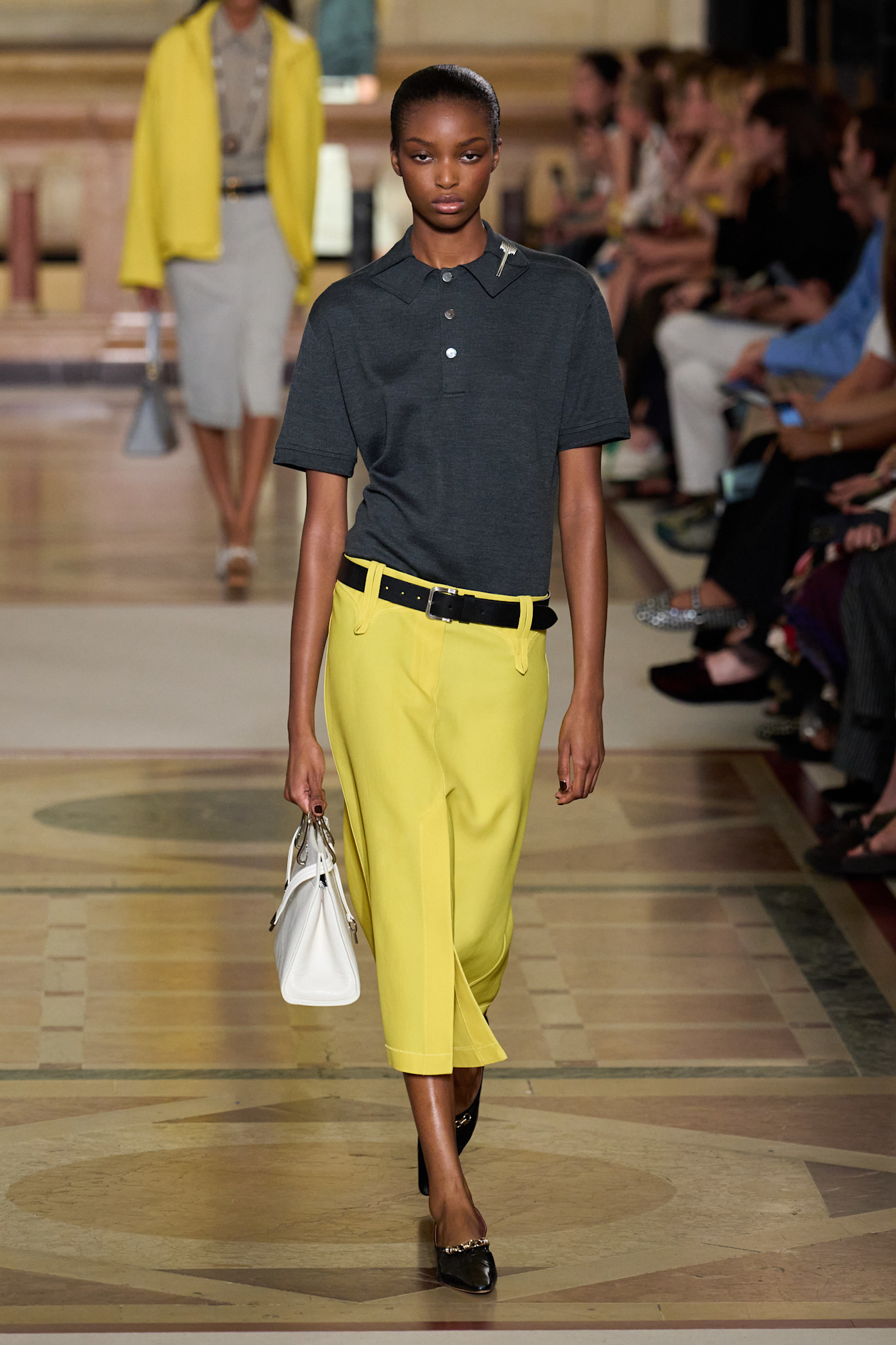 A Tory Burch model wearingya gray polo shirt tucked-into a low-rise citrus yellow pencil skirt styled with a black belt, white top-handle bag and black pointed pumps 