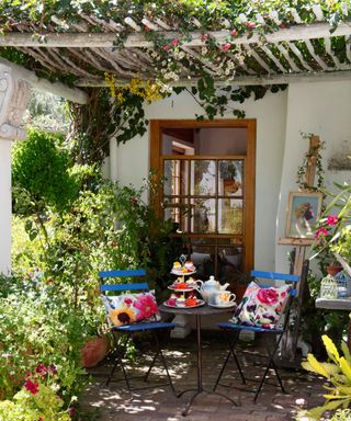 A Mediterranean garden patio with pergola, blue bistro table and chairs with colorful accessories and lush planting.
