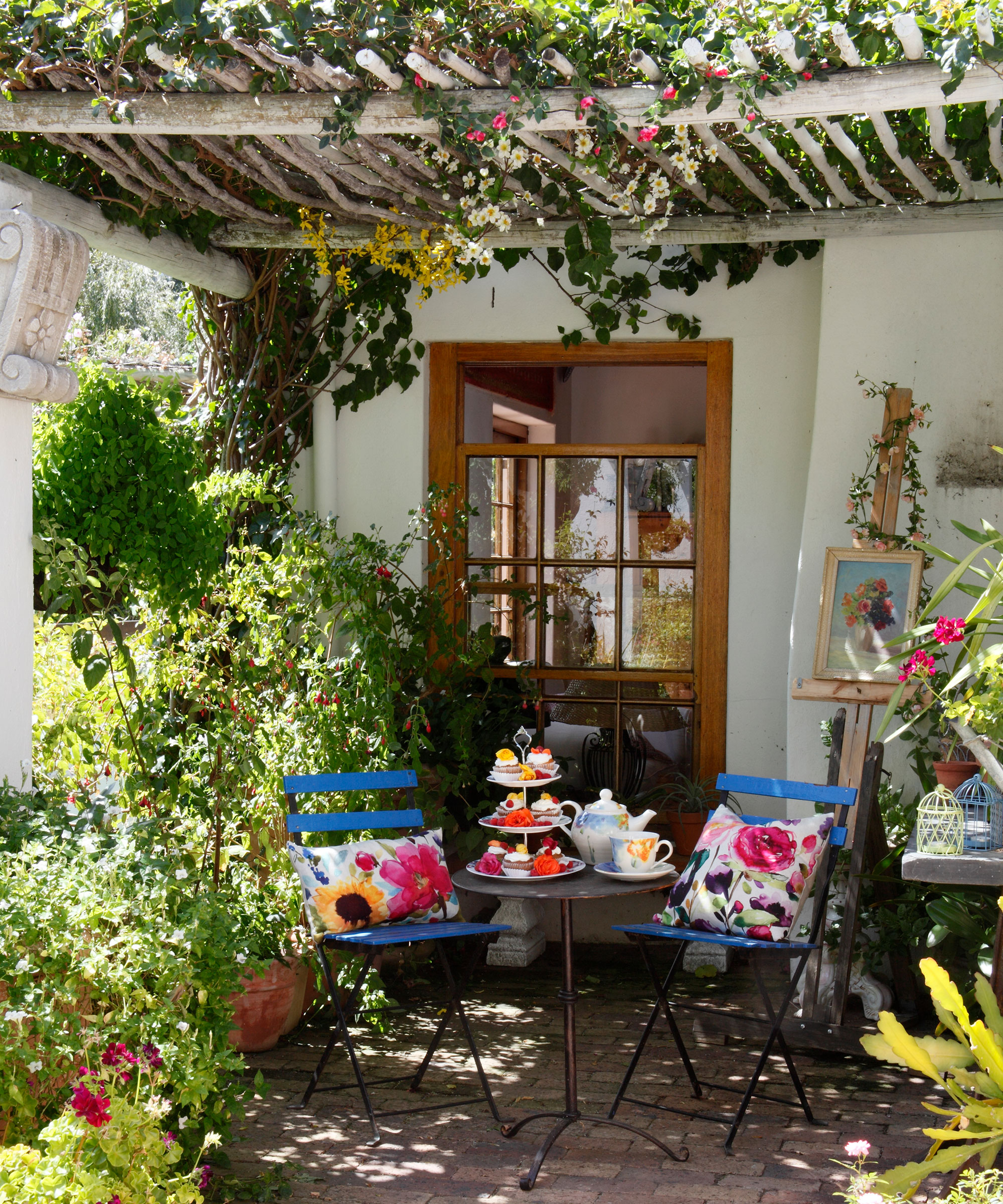 A Mediterranean garden patio with pergola, blue bistro table and chairs with colorful accessories and lush planting.