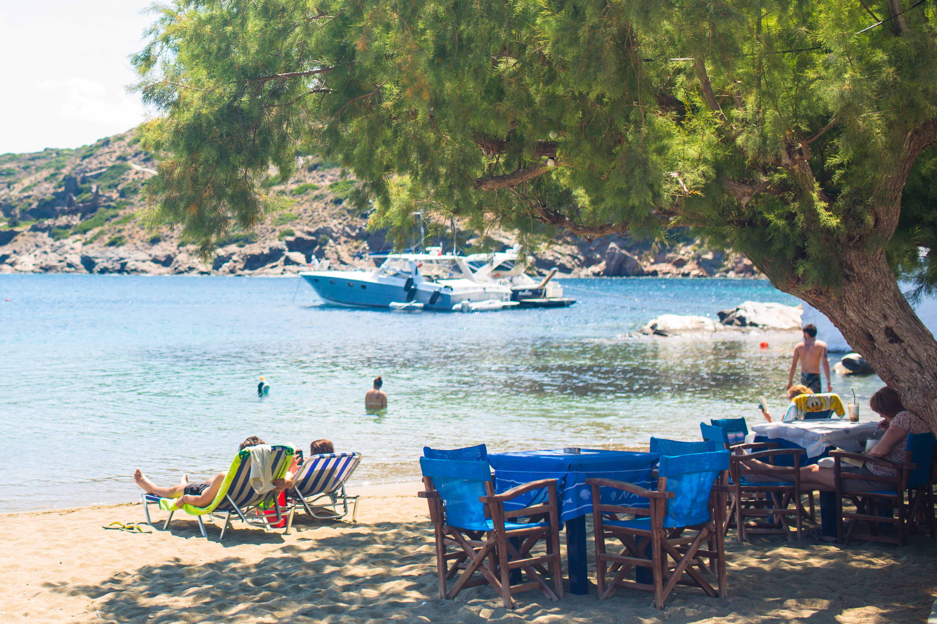 A Greek beach shaded by a large tree. Two people sit in blue and white stripe sun loungers looking out to sea where several boats are moored