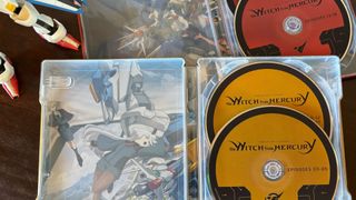 Mobile Suit Gundam: The Witch From Mercury Steelbook discs and cases, on a wooden surface beside an action figure