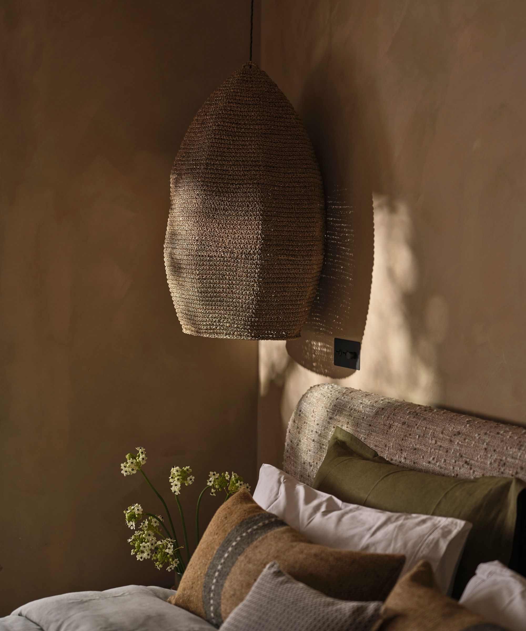 A corner of a neutral bedroom with limewash walls, a long rattan lamp, neutral bedding, and white and green flowers.