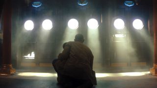 A man kneeling on the floor in a dark room. He's facing several doors that have round windows with light pouring in.