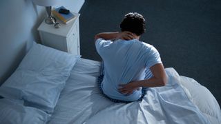 A person sitting on the edge of a bed, holding their neck and back as if in pain