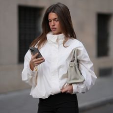 Woman in white high-neck jacket with grey bag looking at her phone. 