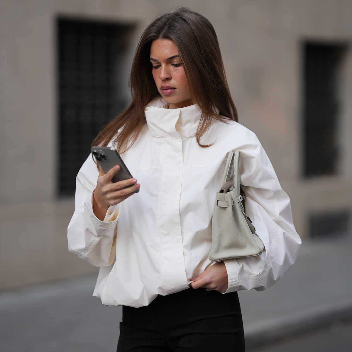 Woman in white high-neck jacket with grey bag looking at her phone. 