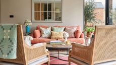 A sitting room with walls in Portland Stone by Little Greene, there is an orange sofa with colorful scatter cushions, and two rattan armchairs with a blue throw. 