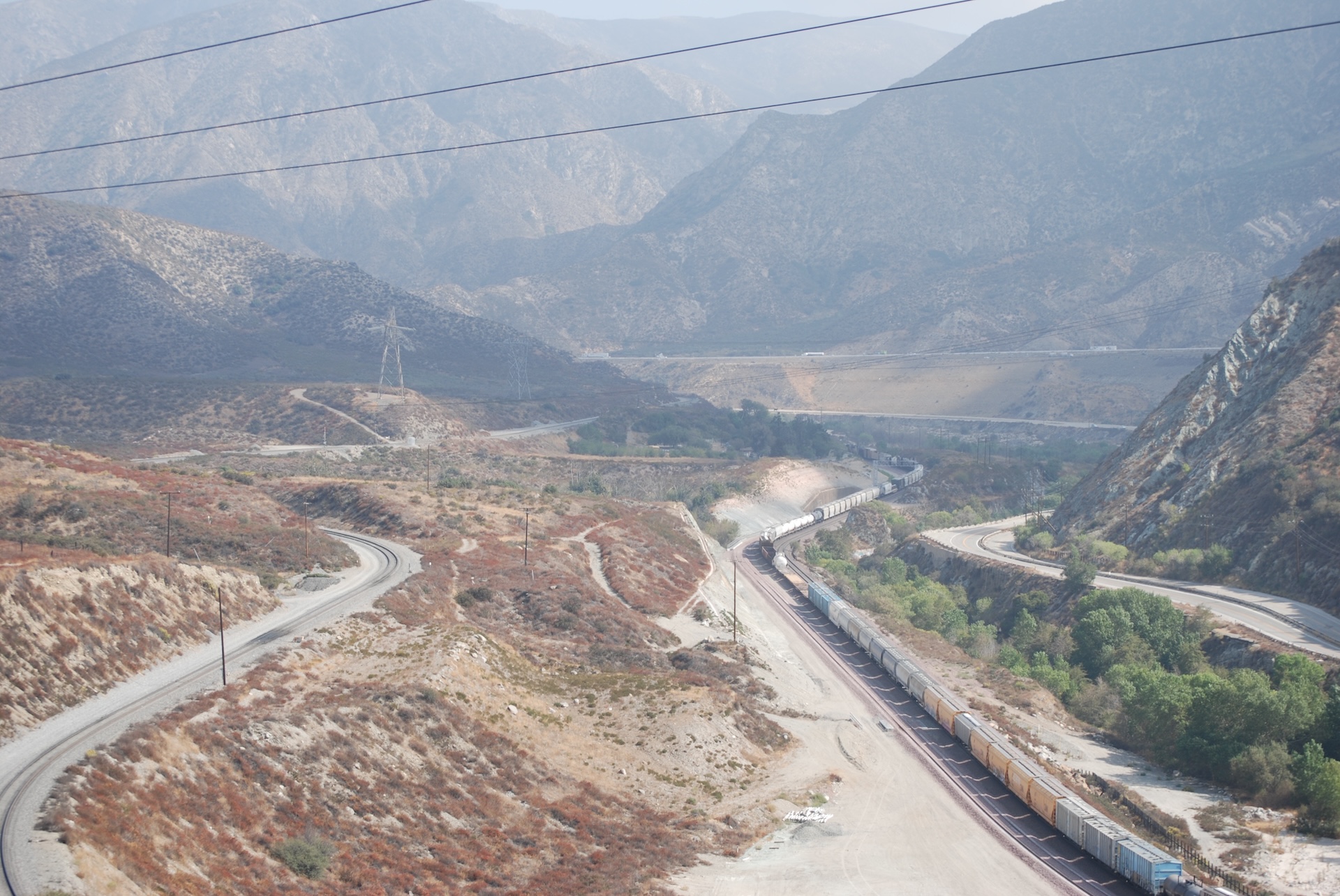 A train track with a freight train runs through a dry mountain valley. Two roads flank the train track.