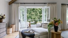 Open balcony window cozy living area of a large bedroom. There is a large sofa lounger with footstool, cabinets with drawers, vases of fresh flowers and long cream curtains framing the background balcony doors