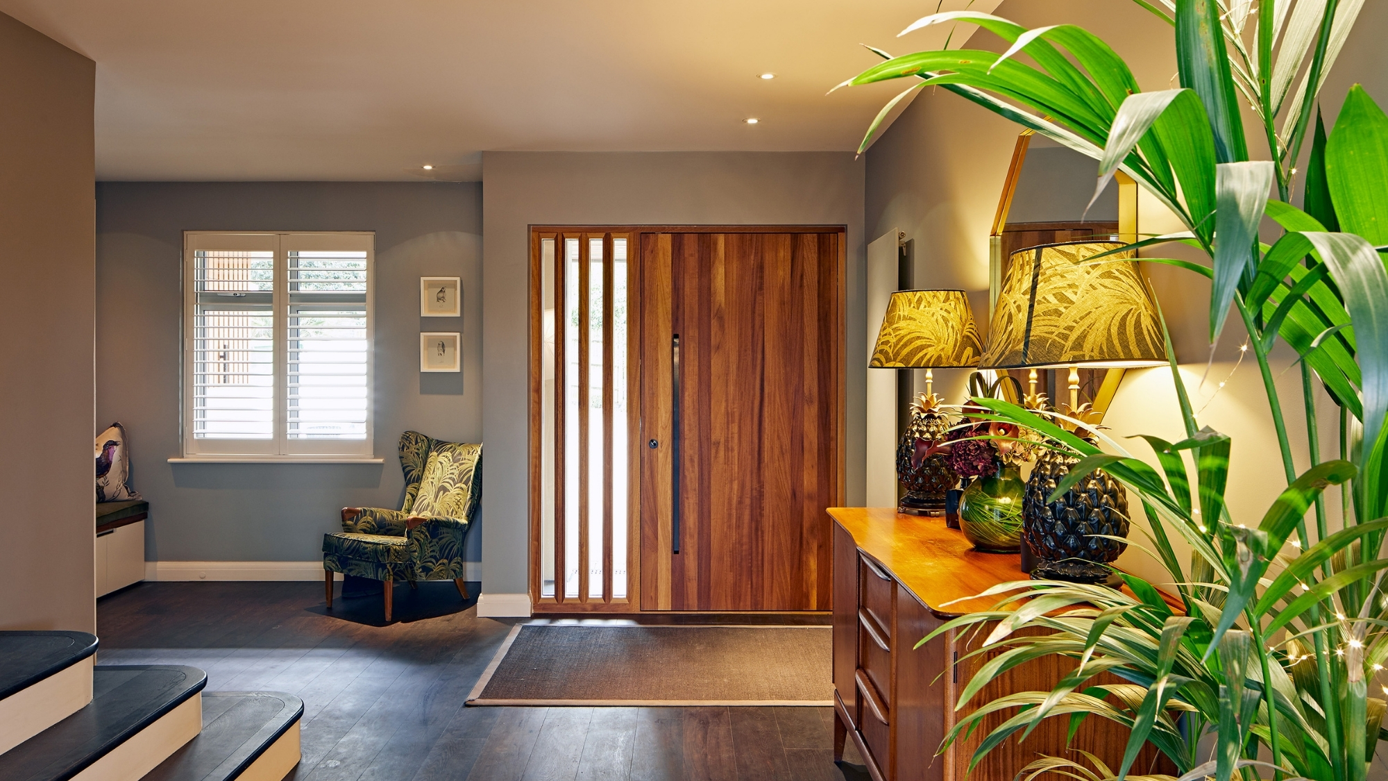 Wooden front door with glass side panels, patterned armchair, sideboard with pineapple lamps and plant.