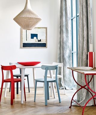 White painted dining room with light wood herringbone flooring, rounded white dining table with black, red, blue and white dining chairs, textured, woven pendant hanging over table, console table with red painted metal frame, wooden tabletop, light gray patterned curtains either side of bright window, blue and white artwork on wall