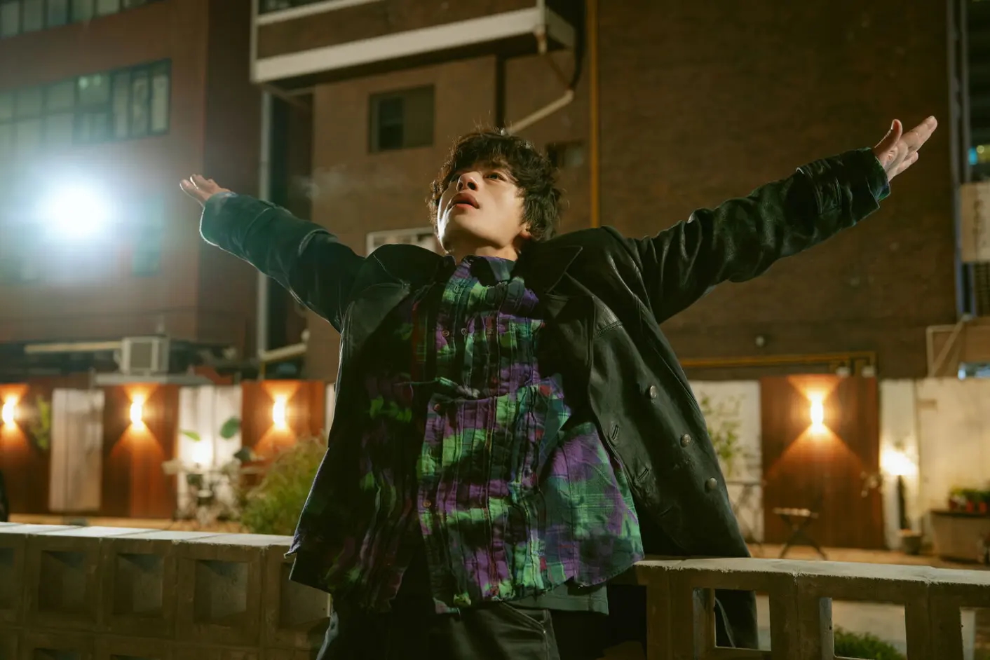 A Korean man (Koo Kyo-hwan) stands with his arms outstretched in front of an apartment building at night, in the K-drama 'We Are All Trying Here.'