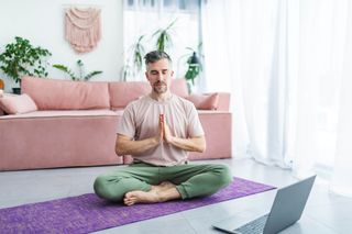 Mid adult man practicing yoga at home online using laptop. Siting in lotus position on exercise mat and meditating with closed eyes