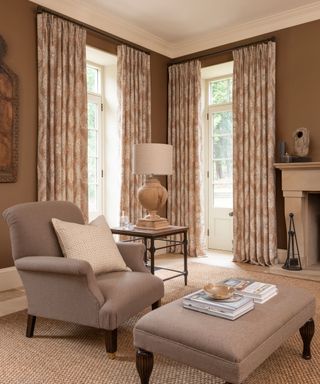 Traditional living room with two large floor to ceiling windows, dressed with cream and brown floral drapes, brown painted walls, light wood flooring, textured natural rug, brown armchair with matching footstool