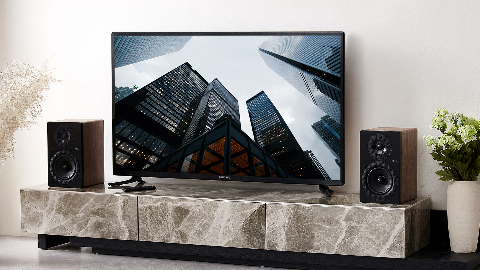 The Elipson Prestige Facet II 6 Active speaker sit on a platform next to a TV, showing they're a compact size that's good for smaller spaces and can replace a soundbar