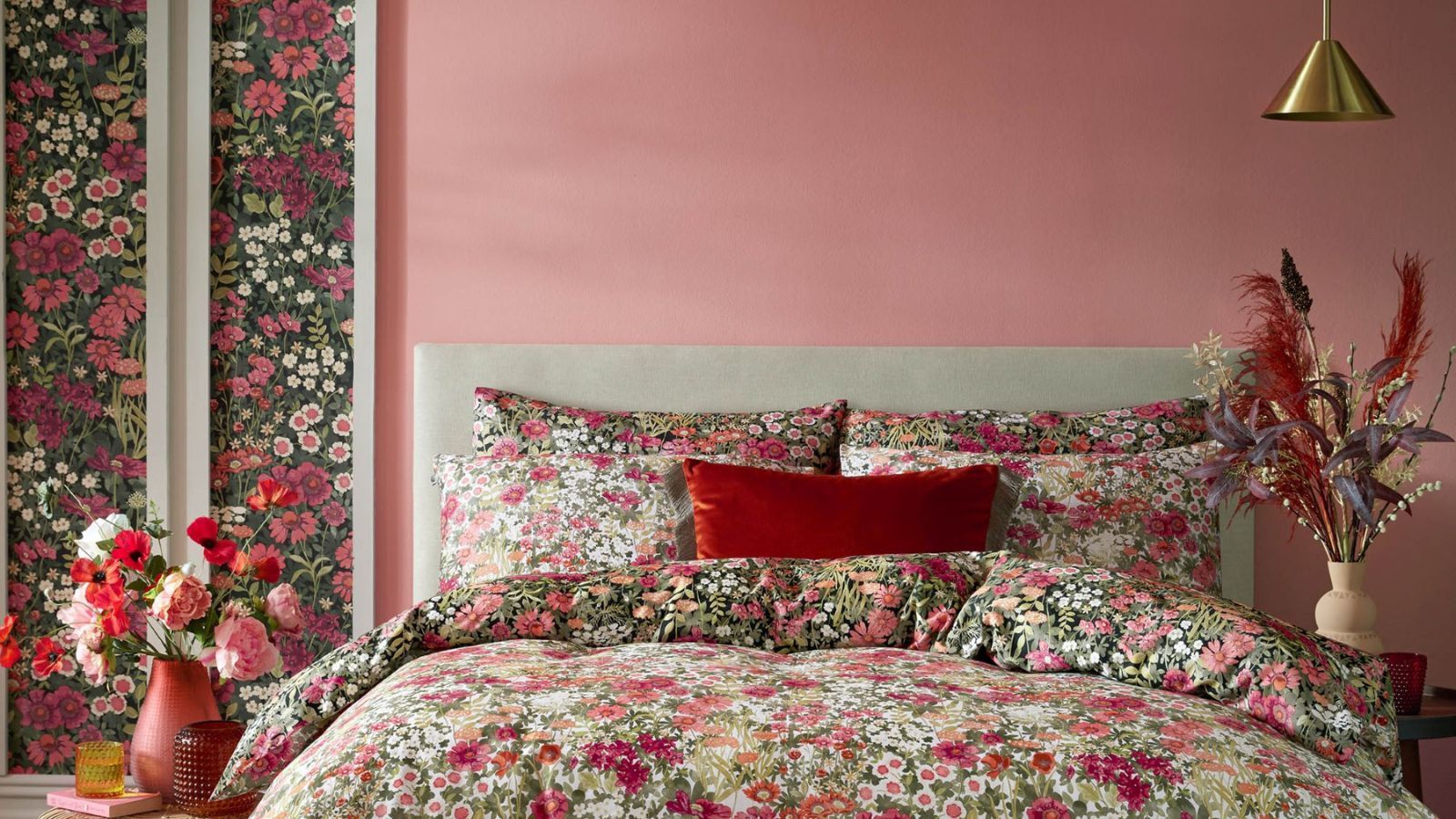 A bedroom with pink and floral walls, a bed with floral bedding and pillows, and vases of flowers either side of it
