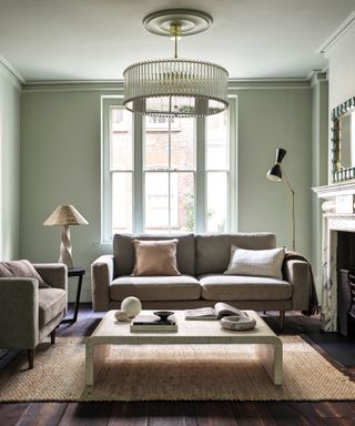 A sage green living room with a glass light, a gray couch, wooden coffee table, and jute rug