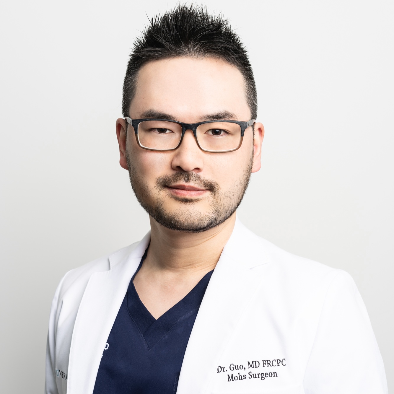 an asian man in dark blue scrubs in a white lab coat with black glasses