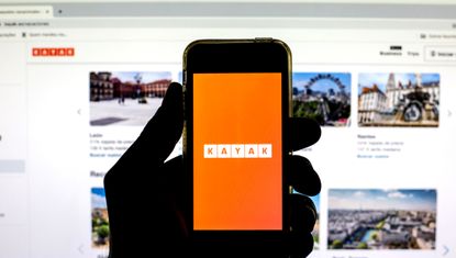 A hand holding a smartphone with the Kayak app displayed on an orange background.