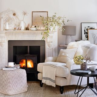 Cosy white living room with white armchair next to a marble fireplace with fire burning
