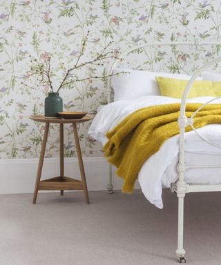 pale carpet in bedroom with white metal bed, floral wallpaper and 3 legged wooden bedside table