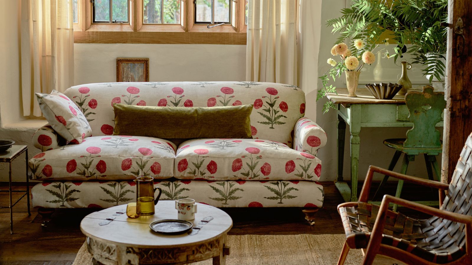 Poppy print sofa in cottage living room. There is a mid-century painted sage wooden sideboard with fresh leaves and flowers on, as well as a mid-century single chair and round distressed small coffee table