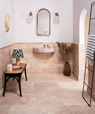 A small bathroom with a pink marble splashback and floor, a pink basin, a rattan side table with a lamp and toiletries on, and a white wall with a curved mirror and two wall sconces